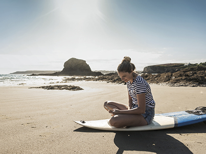 Girl on beach, sitting on surfboard looking at mobile