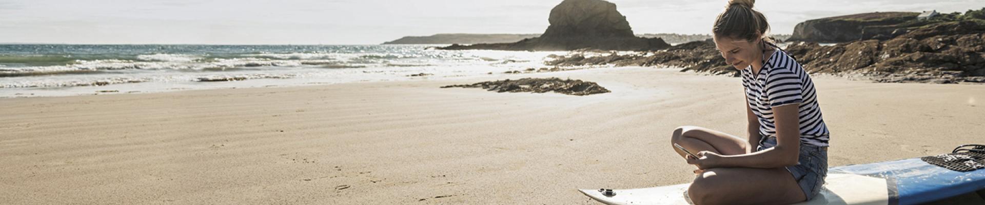 Girl on beach, sitting on surfboard looking at mobile